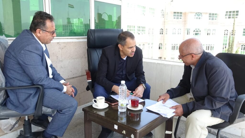 Three men in business attire sit around a small table on a balcony, reviewing documents together.