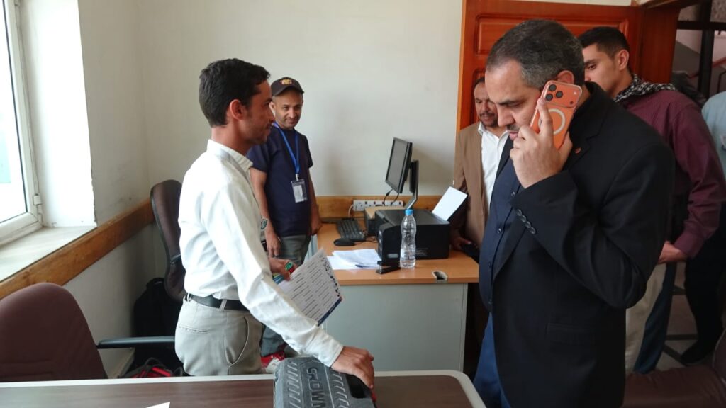 Man in a white shirt hands documents to a man in a dark blazer as colleagues observe in an office, with one person on a phone nearby.