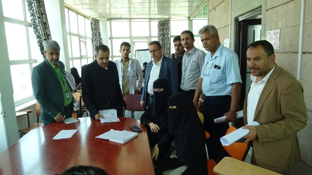 Group of professionals gathered around a large wooden conference table, examining documents in a bright room; two women in black niqab sit among them.
