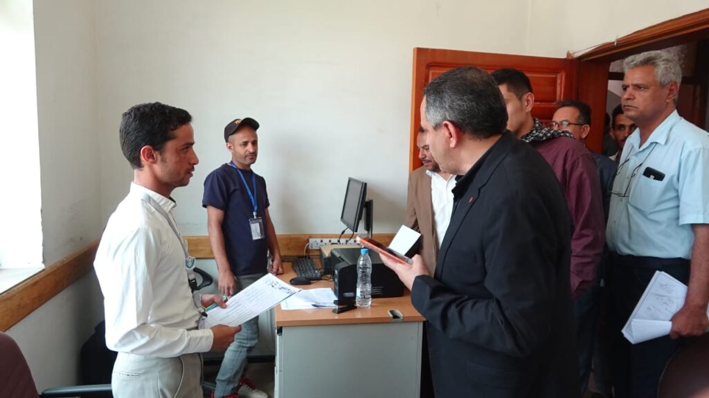 Man in a white shirt hands a document to a man in a black suit in a office, with a computer and water bottle on the desk and several people watching nearby
