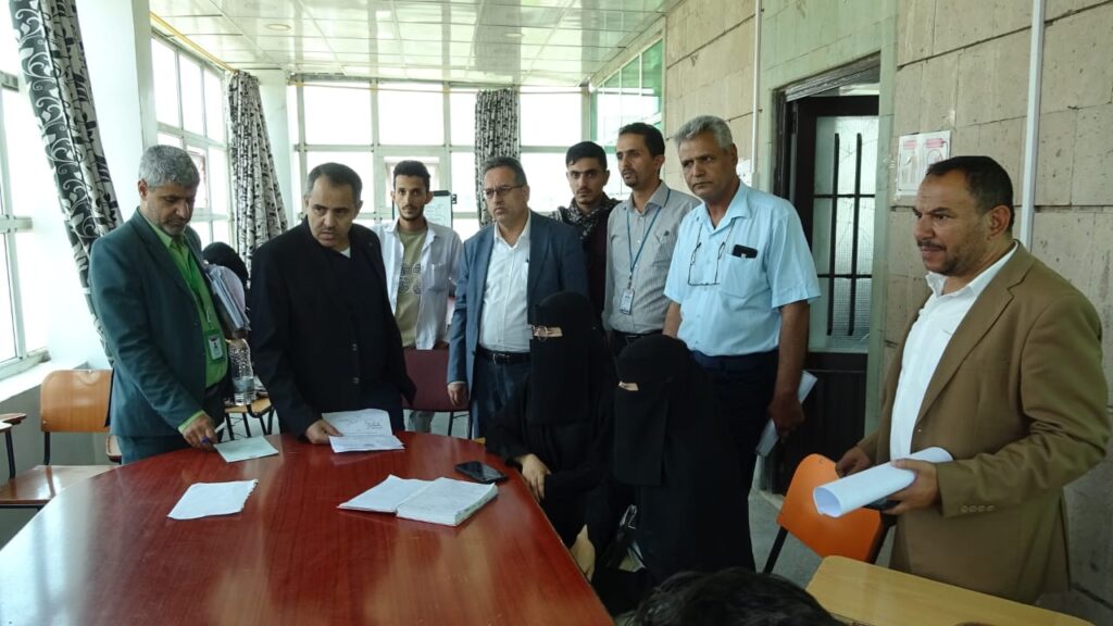 Group of men and two women in niqabs seated around a wooden table with papers in a bright meeting room, discussing documents.