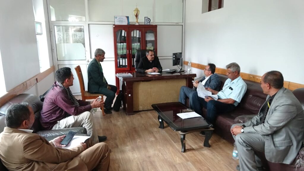 Group of men in a formal office meeting around a wooden desk, some taking notes. (Informative)
