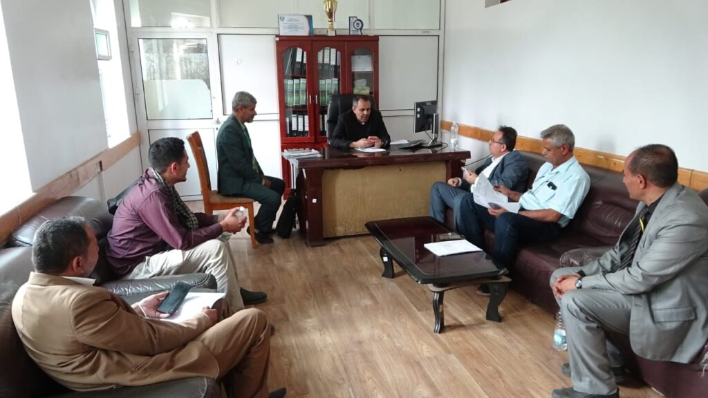 A group of men seated around a office conference area, listening to the person at the desk and reviewing documents.