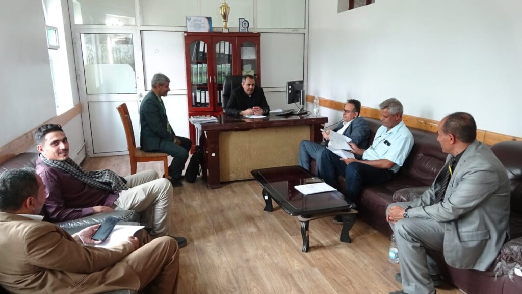 Six men gather in a small office for a meeting: one at a desk with papers, others seated on chairs and a sofa.PCs and documents on the desk; a wooden shelf behind; daylight in.