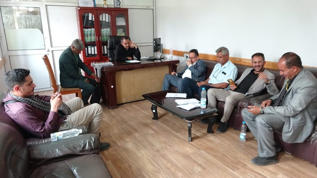 Seven men in an office: one at a desk with papers and a computer while others sit on couches, talking and using phones or tablets.