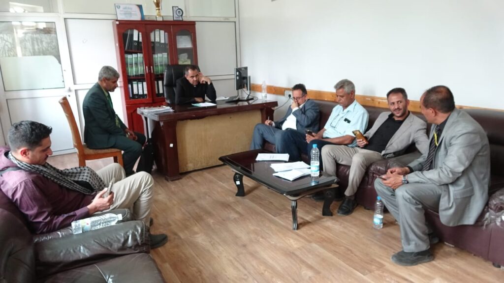 Six men in business attire sit around a desk and on a brown sofa in a small office, discussing with papers and bottles on the table behind them.