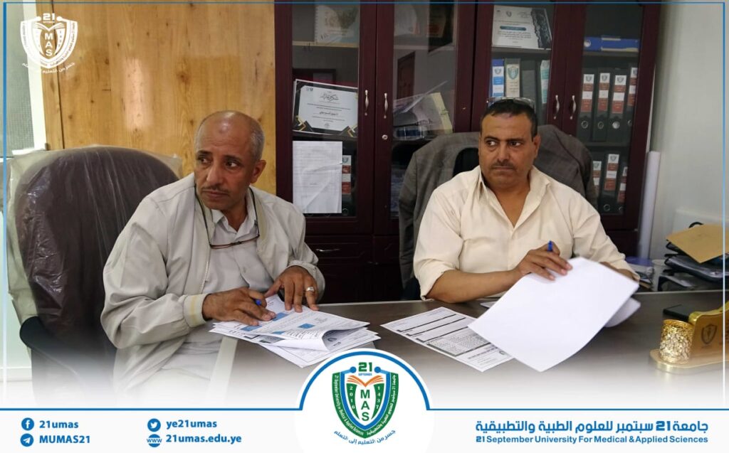 Two men sit at a desk reviewing documents in an office, with the university seal and banner at the bottom edge.