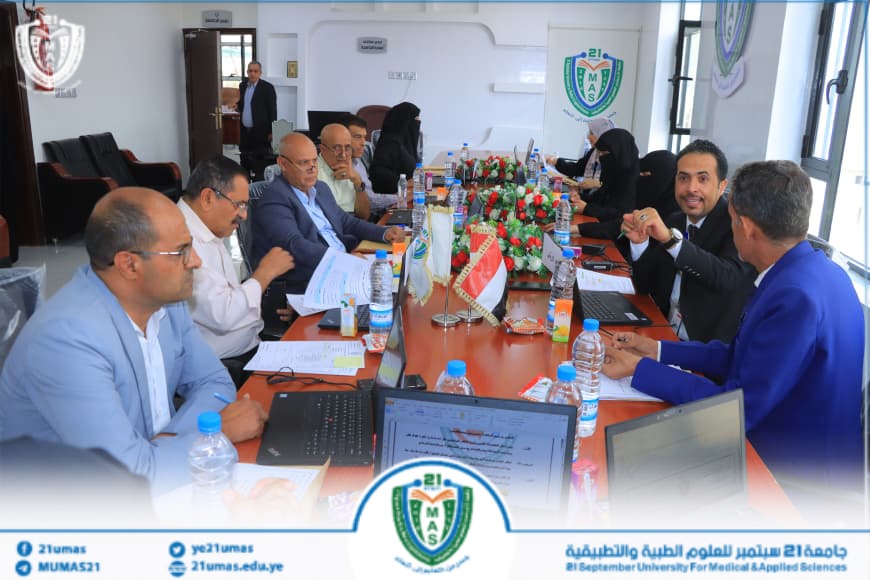 1004048470 - جامعة 21 سبتمبر Group of professionals in business attire seated around a long red conference table with laptops, documents, water bottles, flags, and a university crest on the wall in a bright meeting room.