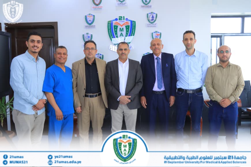 1004048583 - جامعة 21 سبتمبر Seven men in business and medical attire pose for a group photo in a university office, with crest logos and banners on the wall behind them.