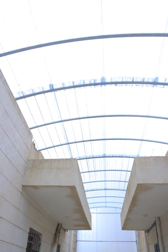 Narrow beige concrete walkway between buildings under a translucent curved roof with metal ribs.