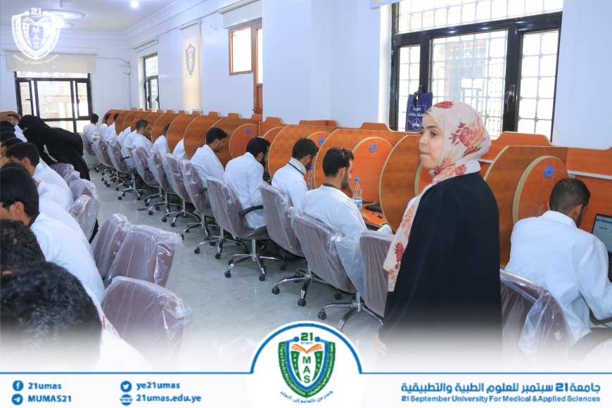 Students in white shirts seated at computer desks with orange privacy dividers in a computer lab; a woman wearing a floral hijab stands in the foreground, overseeing the room.