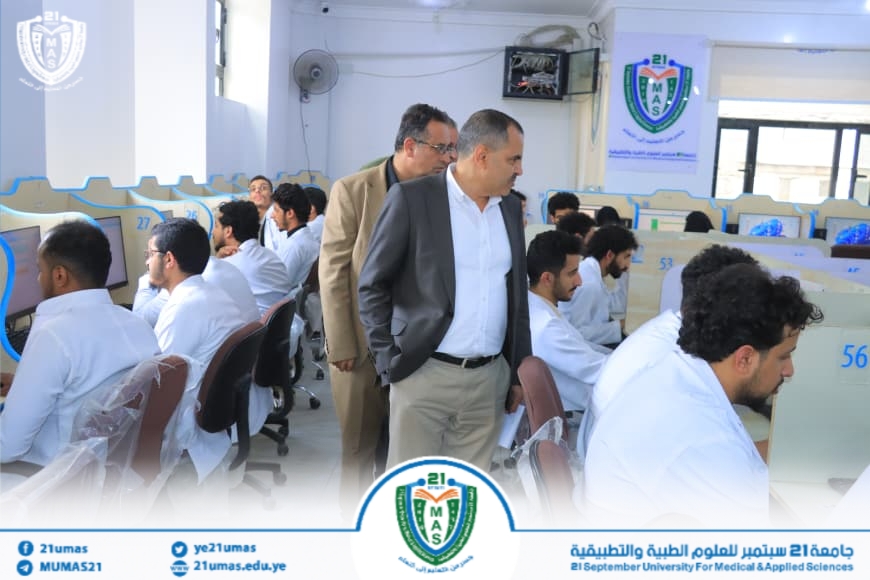 Group of men in business attire walk through a computer lab filled with students in white lab coats seated at cubicles; university crest and banners are visible.