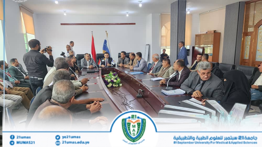 Group of officials seated around a long conference table in a formal meeting room; two flags behind them and reporters filming on the side, banner with the university logo at the front.