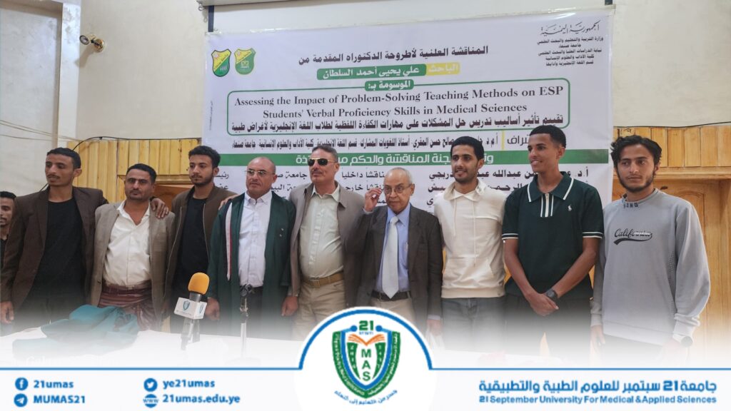 1004053933 - جامعة 21 سبتمبر Group of men in smart casual attire posing together on a stage in front of a large banner with Arabic and English text about a university event; microphones and a desk are visible in the foreground.