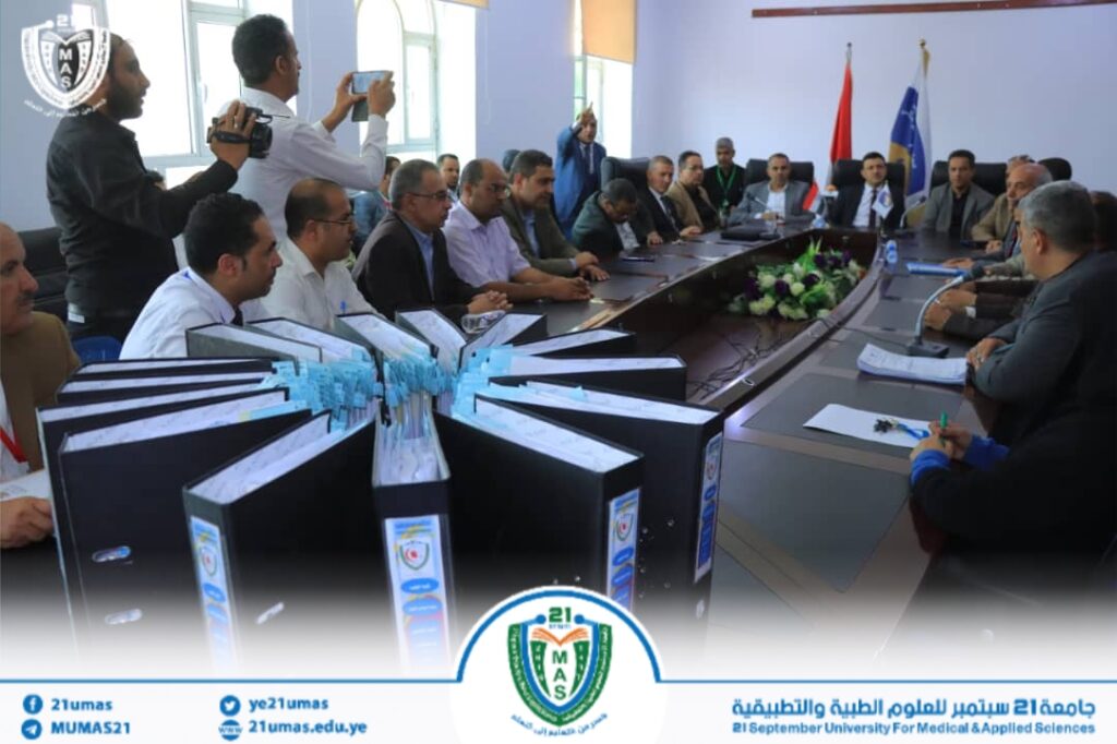 Formal meeting in a university boardroom with many attendees around a long conference table; a photographer documents the event, banners and the university crest visible on screen.
