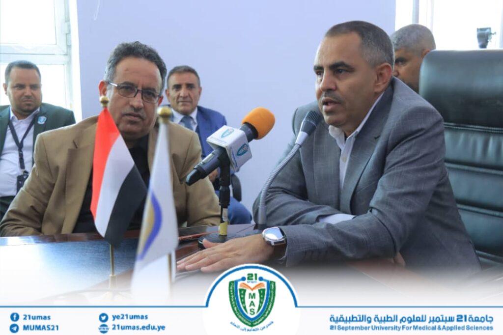 Two men in suits speak at a press conference with microphones and flags on the table, banner and university logo visible in the foreground.