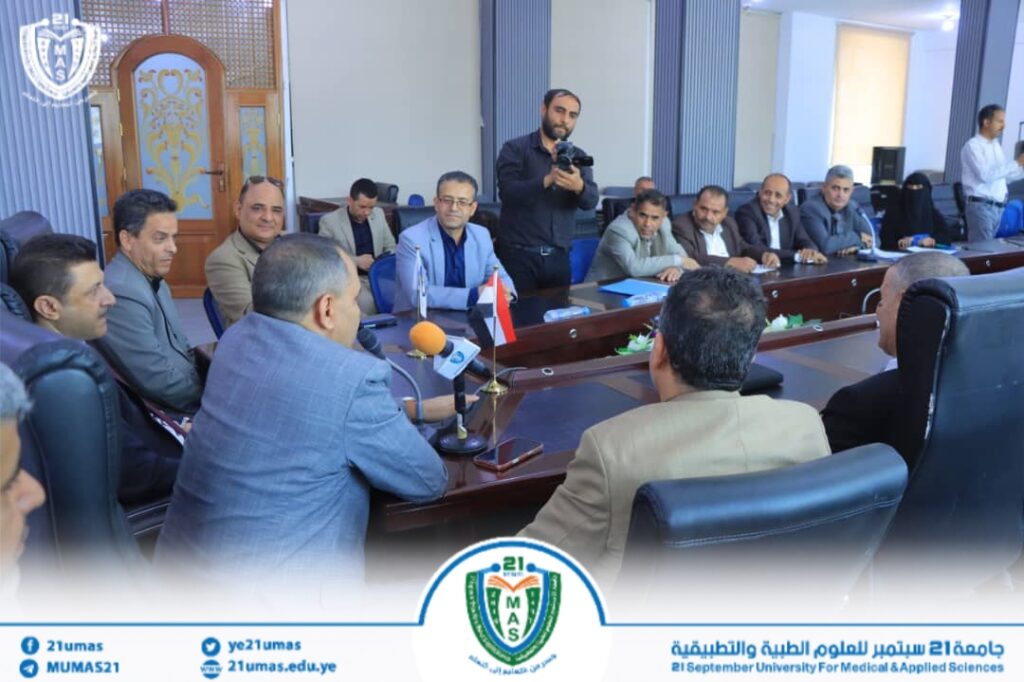 Group of professionals around a long conference table; a photographer stands in the background, microphones on the table, university banner visible at the bottom.