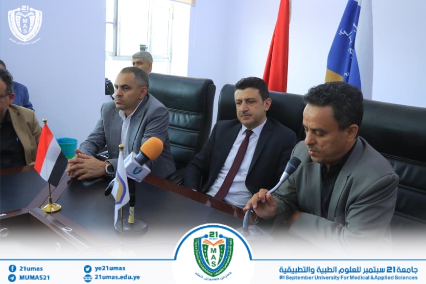 Three men in suits sit at a conference table with microphones during a press briefing at 21 September University. Flags and a crest are visible on the banner backdrop.