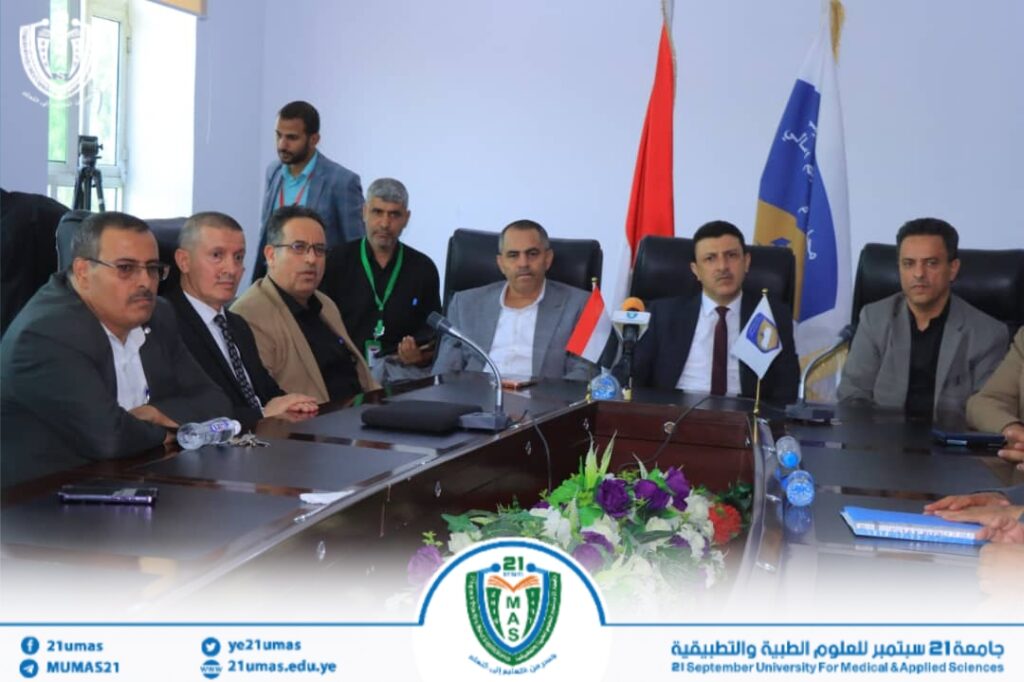 Group of men in formal attire seated around a conference table during a meeting at 21 September University for Medical & Applied Sciences, with flags and flowers in the foreground.