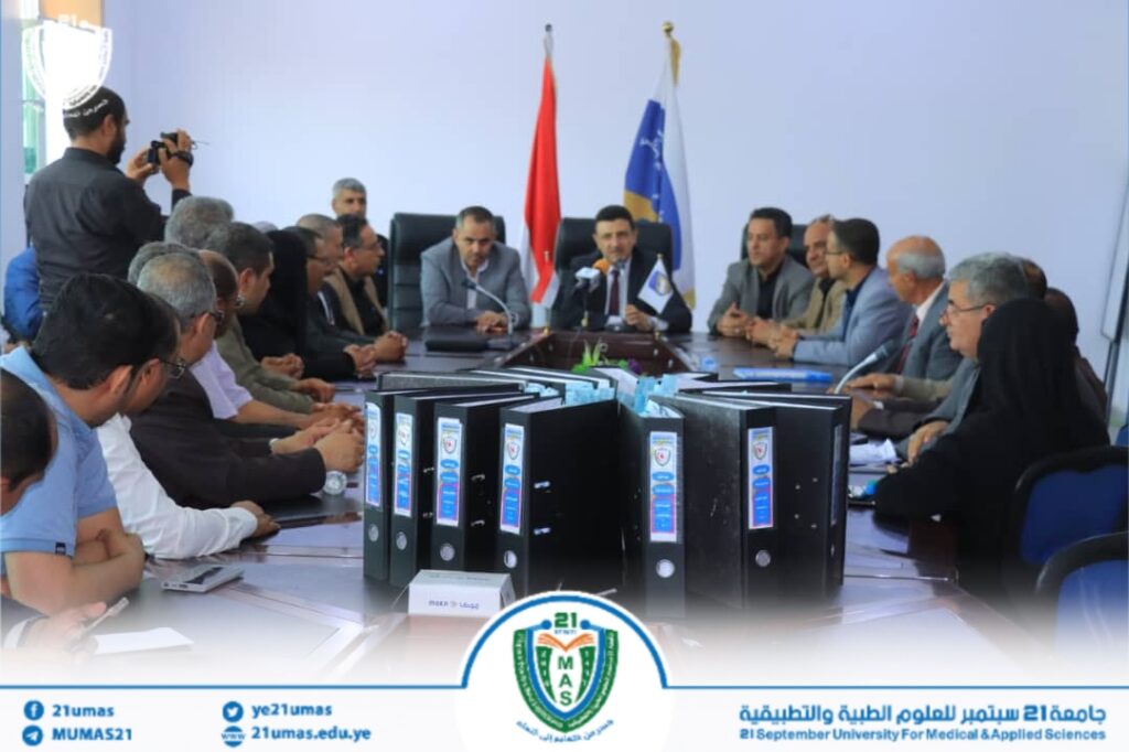 Group of university officials seated around a long conference table for a formal meeting, binders in the foreground and flags in the back.