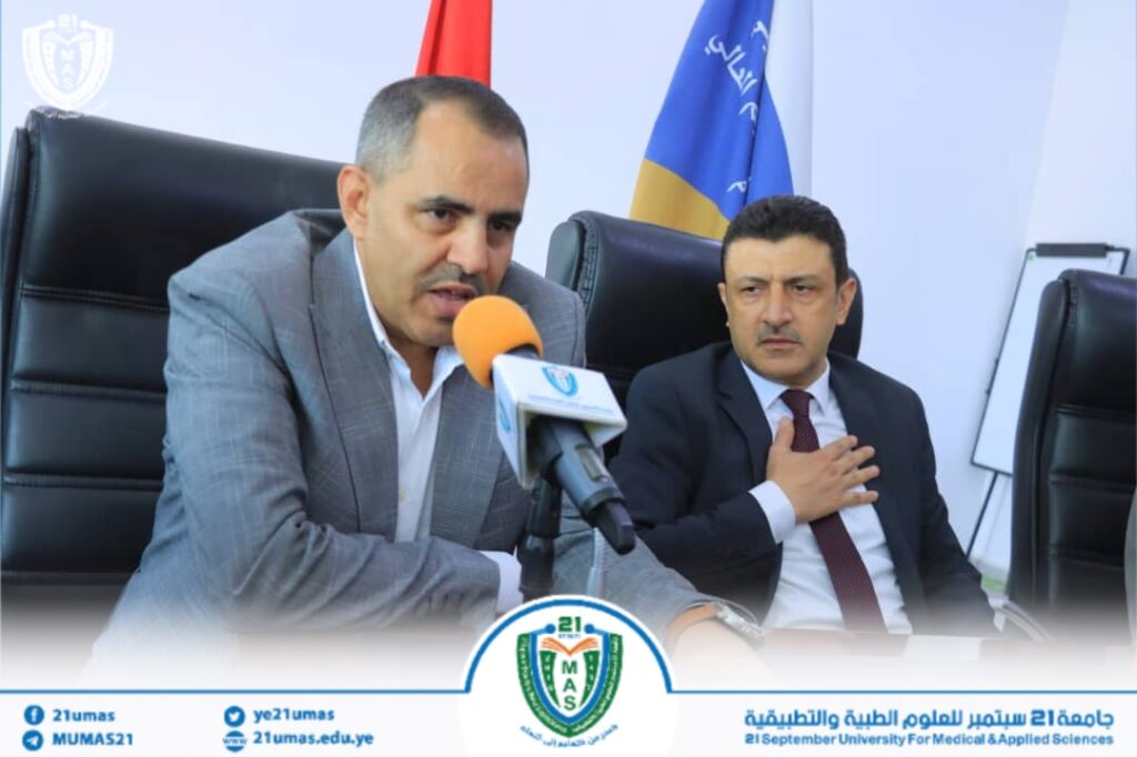 Two men in suits seated at a press conference with a microphone in front of them, university logos visible on the banner behind and at the table front.