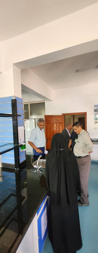 A woman in a black abaya stands at a service counter while three men discuss at the opposite side in a public building.