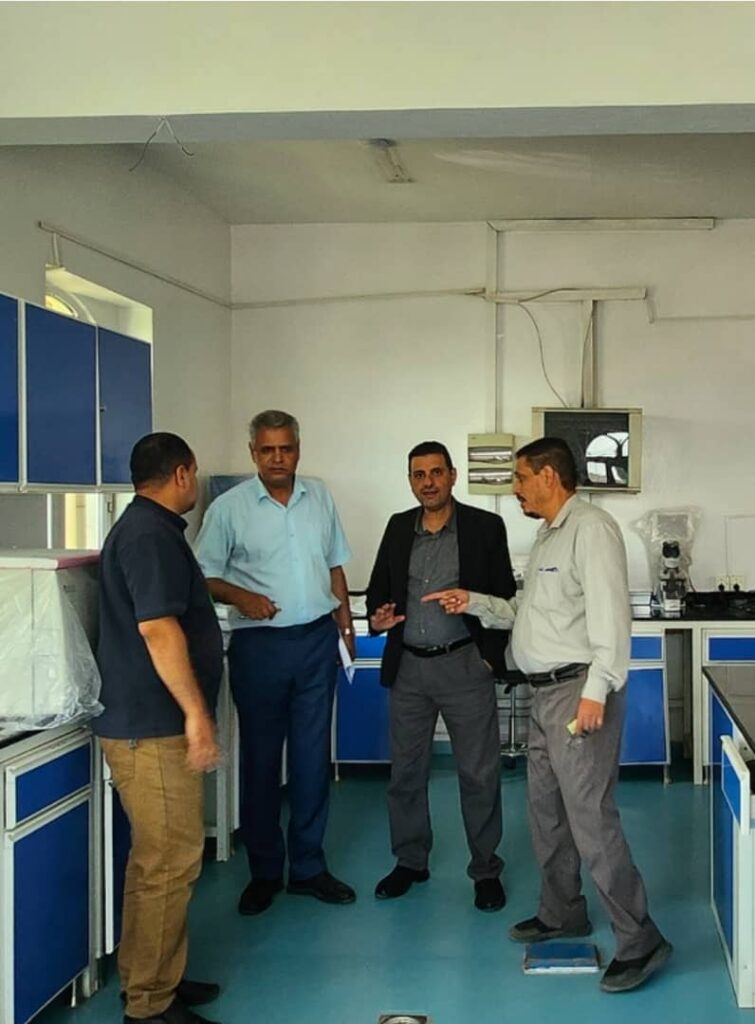 Four men in a science lab standing near blue cabinets, engaged in conversation around a workstation.