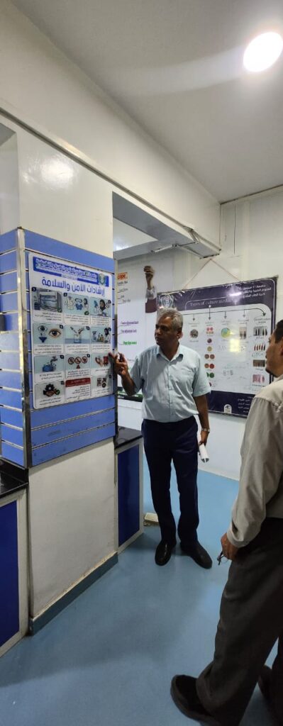 A man in a light blue shirt points at an informative poster in a classroom or health station setting as another person looks on.