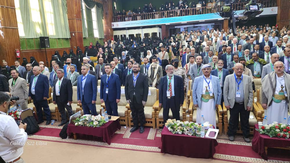 Large crowd of men and women in formal attire standing and seated in a conference hall, facing the stage.
