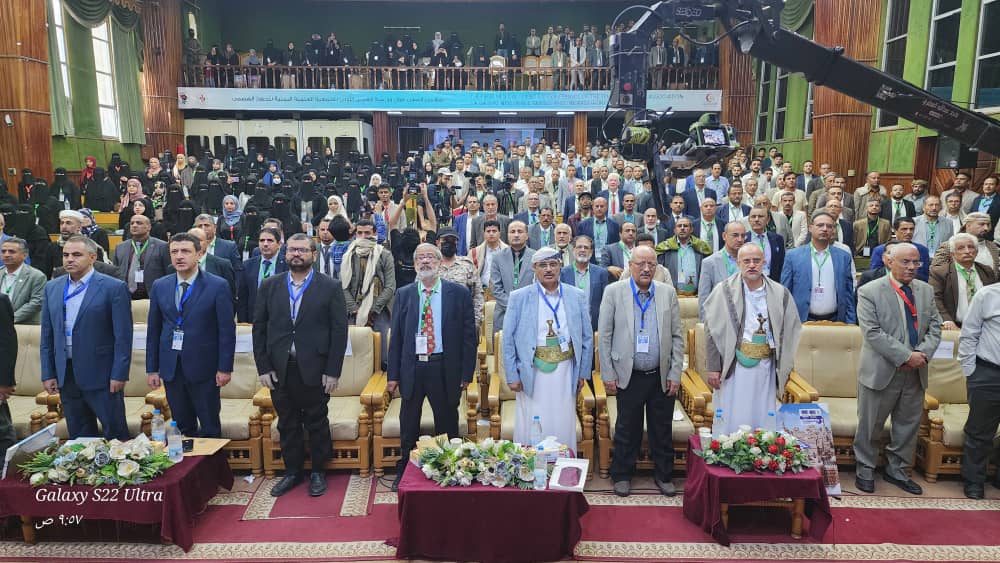 Large group of conference attendees standing in a hall, front row in suits and traditional garments with bouquets on tables in front.