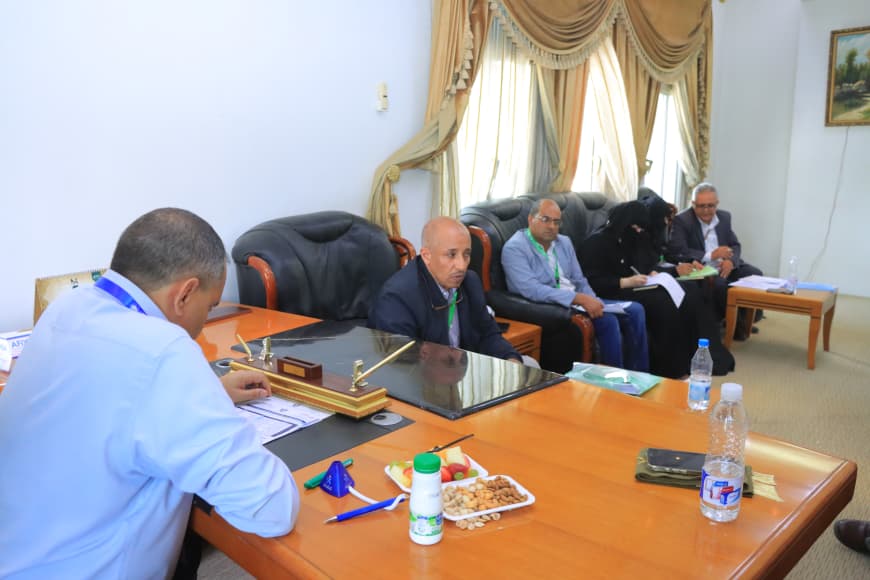 Man in a light blue shirt sits at a large wooden conference table taking notes; in the background, five people sit on a leather sofa, with snacks and water on the table