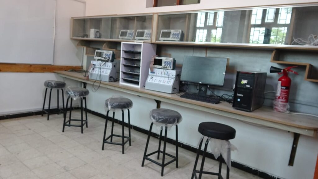 Electronics lab workstation with multiple testing instruments, a computer, shelves, and several stool chairs, some covered in plastic, along a long counter. A fire extinguisher is on the far right.