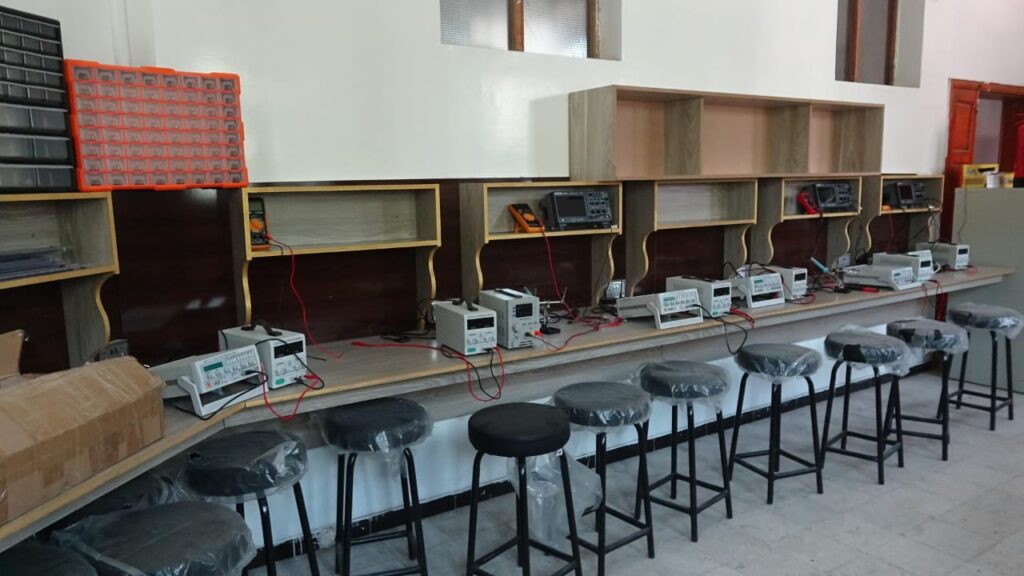 Electronics lab bench with multiple power supplies and oscilloscopes, red test leads, and stools in plastic wrapping.