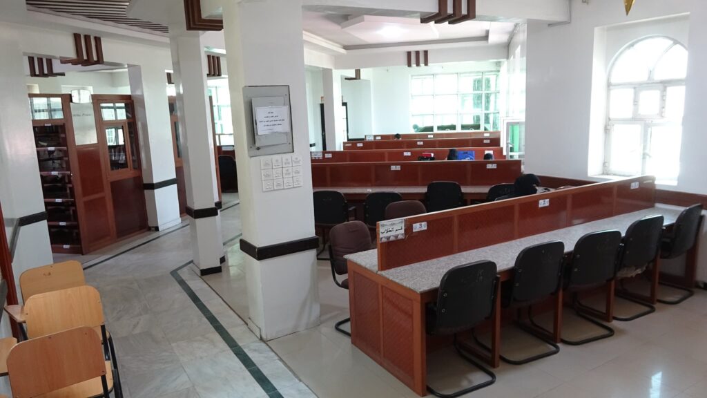 Study hall or library with long rows of brown partitioned desks and black chairs, white columns, and large windows at the far end.