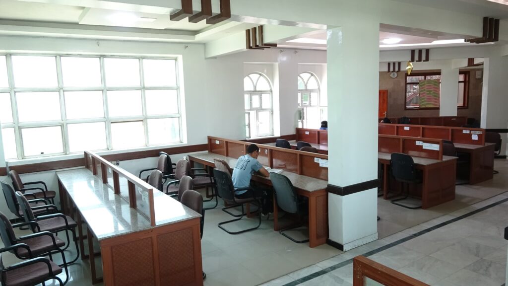 A spacious study hall with long wooden desks and many chairs, large windows on the left, and a student in a blue shirt seated at a desk in the middle-right area.