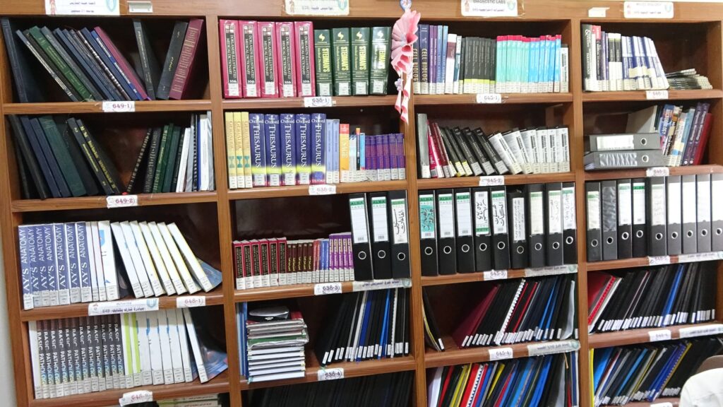 Rows of medical textbooks on a wooden library shelf, with call-number labels and a pink decorative flower in the center.]