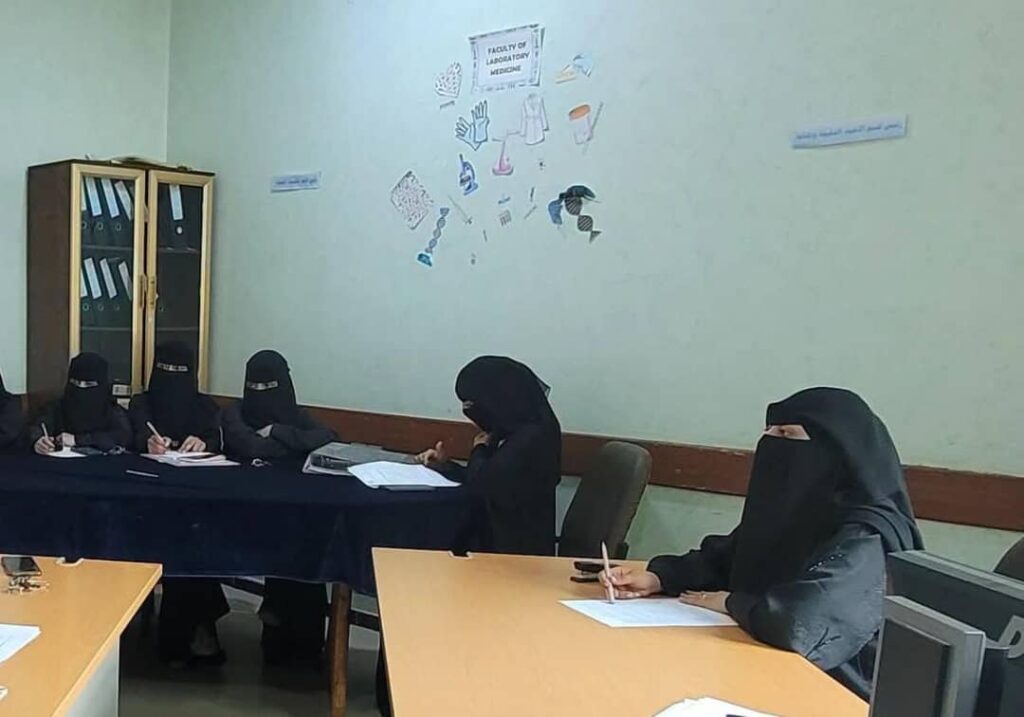 Five women wearing niqabs sit around a table, taking notes in a classroom.