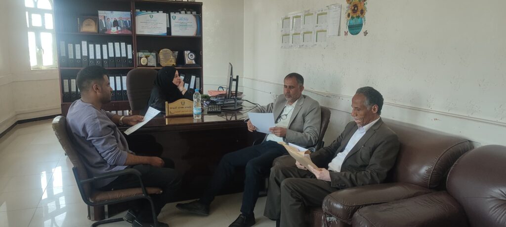 Four professionals sit around a desk in an office, reviewing documents during a meeting (woman in hijab at center).