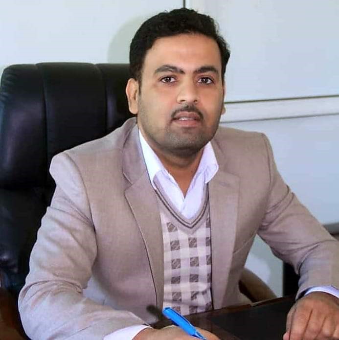 العميد - جامعة 21 سبتمبر Man in a beige blazer seated at a desk in an office, holding a blue pen and looking at the camera.