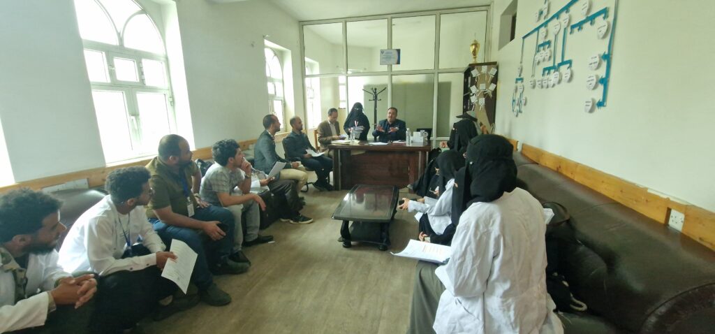 Group of people in a meeting room, seated in a semicircle around a wooden desk while a presenter explains documents.