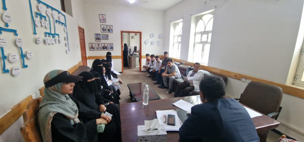 Group of people seated around a long conference table in a small meeting room; women in black hijabs sit on the left, men on the right, with portraits on the wall behind them.