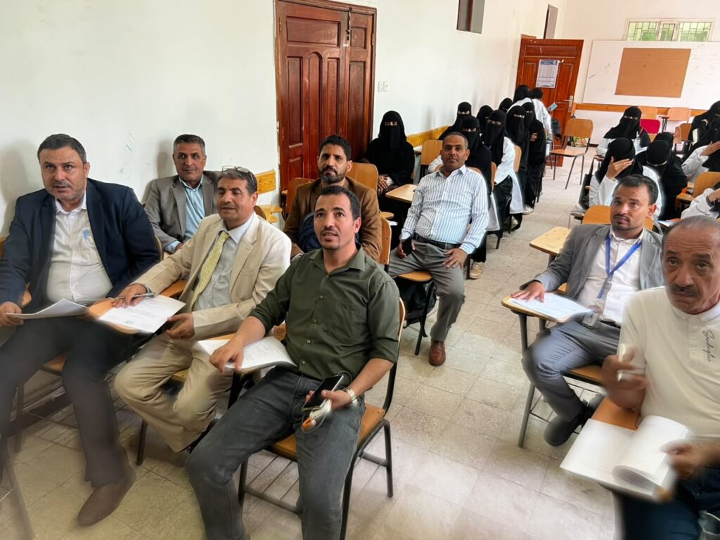 Group of men sits in a classroom with notebooks, while women in black hijabs sit in the back rows.