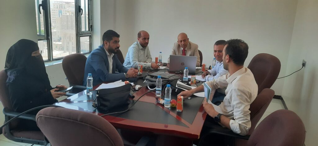 Six professionals seated around a conference table with laptops and documents during a meeting; a woman in a black hijab sits at the left.