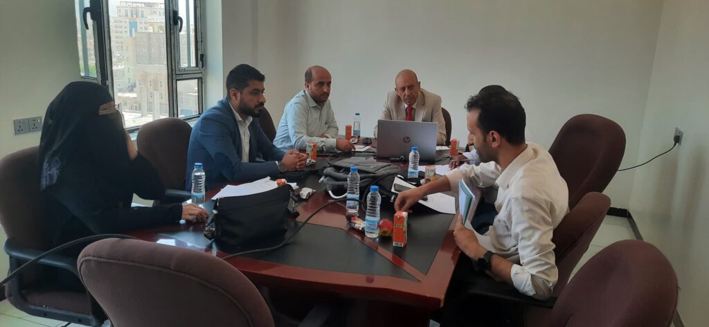 A group of people seated around a conference table in a bright office, discussing with laptops, papers, and drinks on the table.