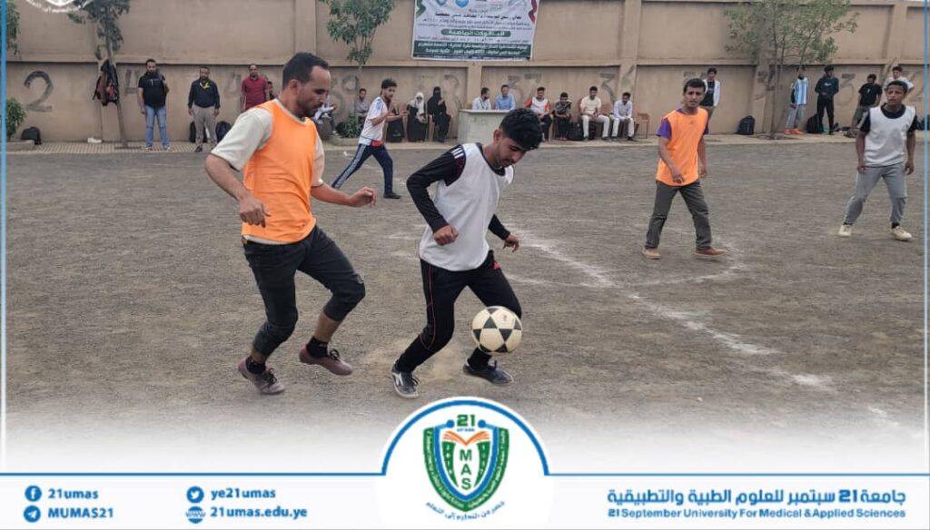 Young men play soccer on a dusty outdoor field as one player controls the ball while another pursues in an orange bib. Spectators stand along a wall in the background, with banners visible above.