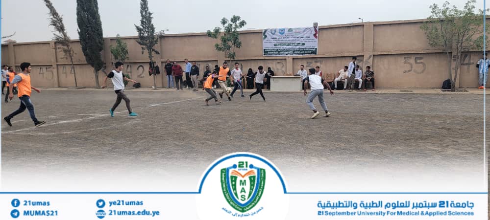 Group of students playing soccer on a dusty outdoor field, some in orange bibs, with a wall and banner in the background and spectators along the wall; university logo at the bottom.