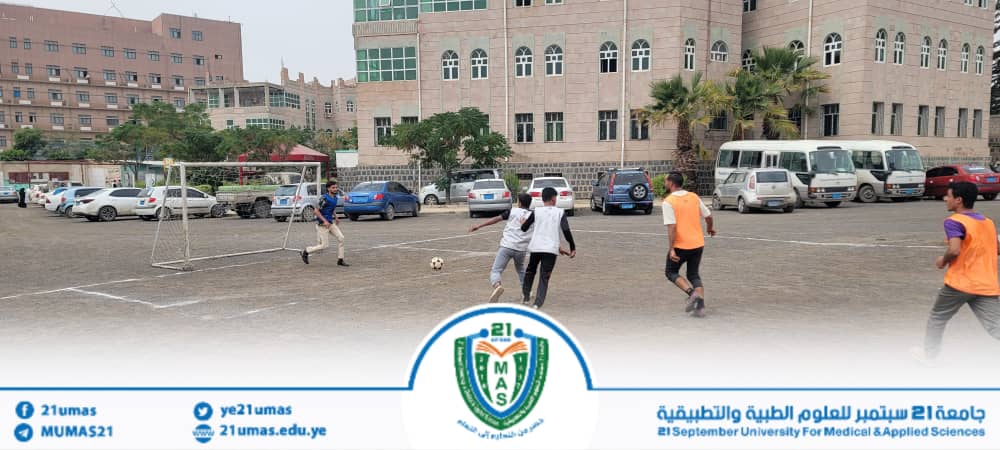 Group of students playing soccer on a campus field, with a goal and parked cars in the background and a large building behind them.