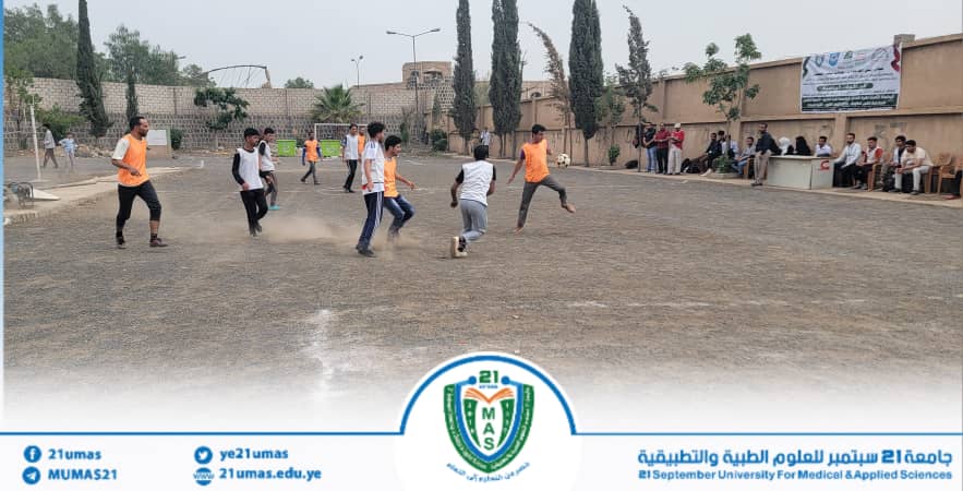 Students play a casual soccer match on a dusty outdoor field, with a banner and spectators along the sidelines behind them.