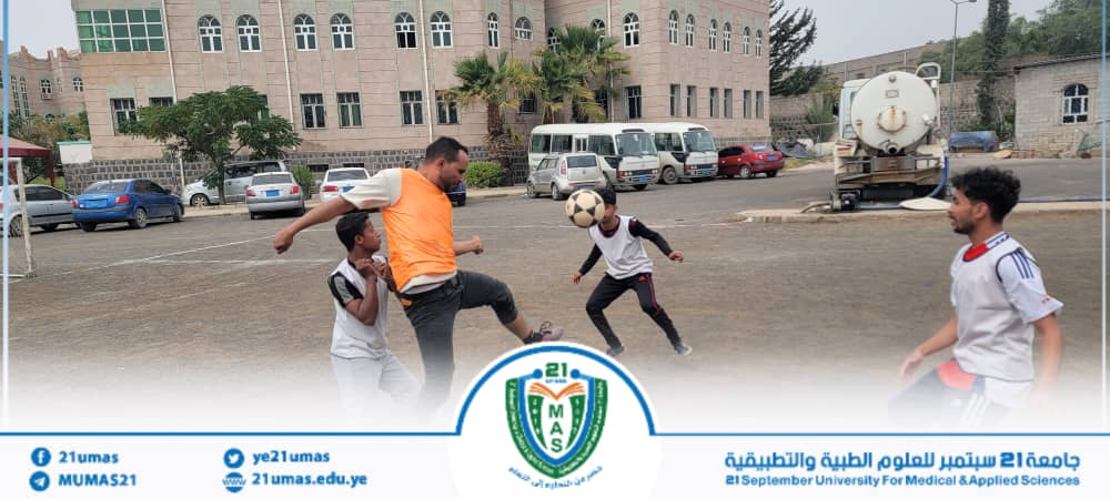 A man in an orange shirt kicks a soccer ball while kids play nearby near a campus building; a university banner with logo bands the bottom edge.