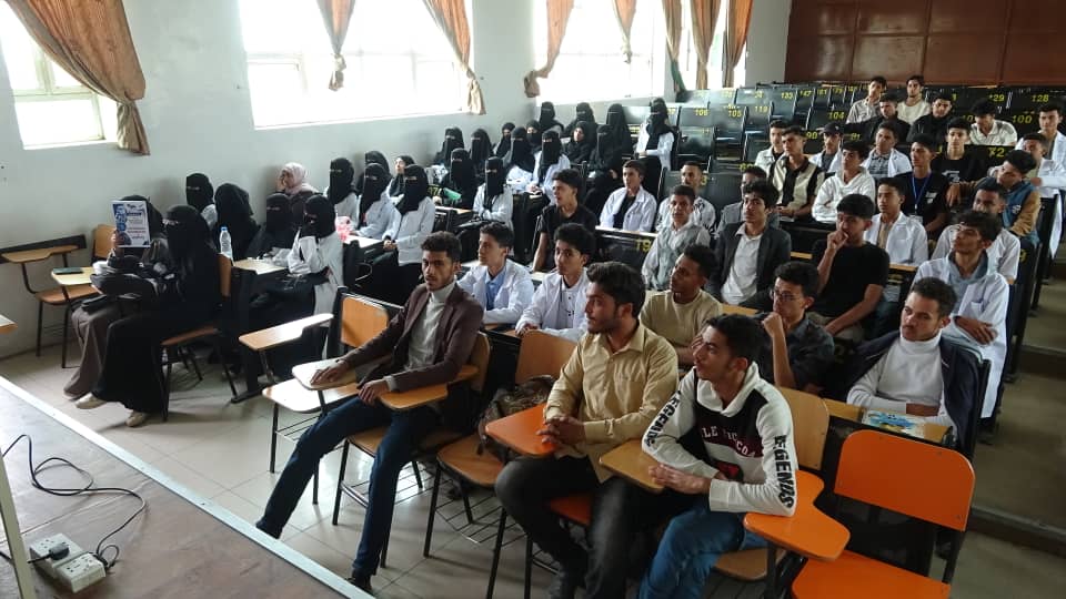 Lecture hall filled with students in orange chairs, many women wearing hijabs or niqabs, listening to a lecture at the front of the room.